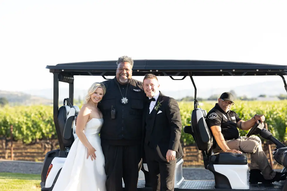 Carl Vinson in a security vest walks beside a bride and groom on a golf cart at a vineyard wedding while a second guard drives